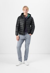 Zwarte pufferjack met capuchon, voorzien van een groene binnenvoering, ritssluiting en verticale quiltstiksels. Geddragen over een donker shirt en lichte spijkerbroek.