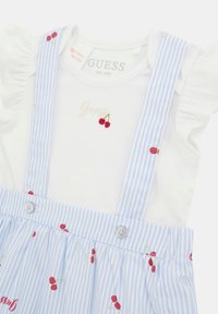 Camicia bianca a maniche corte con maniche arricciate e design di ciliegie ricamate, abbinata a una salopette a righe blu con accenti rossi a forma di ciliegia.