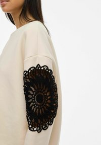 Beige sweatshirt featuring a black lace crochet accent on the left sleeve, with a floral pattern and scalloped edges.