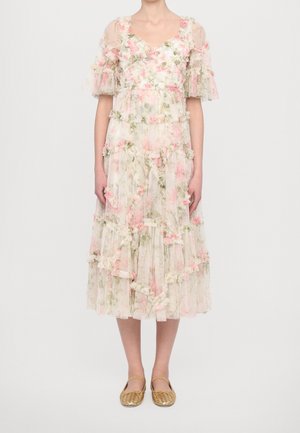 Vrouw die een lichte midi-jurk met bloemenprint draagt, met geplooide lagen en doorschijnende korte mouwen, gecombineerd met gouden gevlochten platte schoenen.