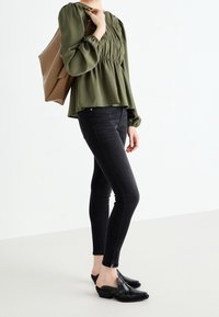 Haut vert olive à manches bouffantes avec détail froncé, porté avec un jean skinny noir et des mules à talons noires avec des coutures décoratives. Sac cabas marron.