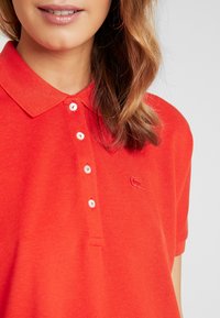 Femme portant un polo rouge avec quatre boutons blancs et un logo de crocodile brodé sur la poitrine gauche, visage et cheveux partiellement visibles.