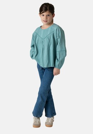 Niña joven de pie con las piernas cruzadas, vistiendo una blusa de manga larga texturizada en color verde azulado, vaqueros azules y zapatillas de color claro.