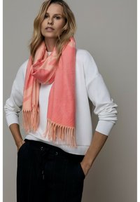 Écharpe rose à texture douce, avec des bords effilochés. Portée sur un sweat-shirt blanc, contrastant avec un pantalon foncé. Design simple et élégant.