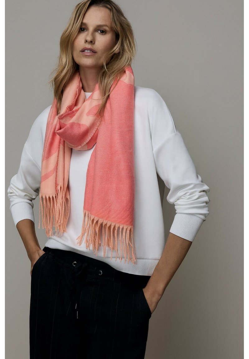 Écharpe rose à texture douce, avec des bords effilochés. Portée sur un sweat-shirt blanc, contrastant avec un pantalon foncé. Design simple et élégant.