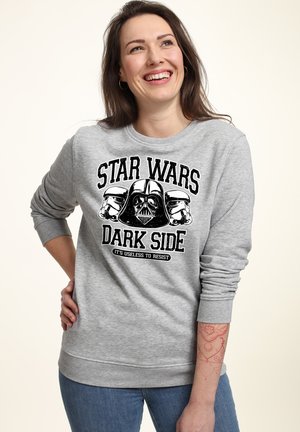 Smilende kvinde med langt mørkt hår iført en grå Star Wars sweatshirt med Darth Vader og stormtroopers, stående med hånden på hoften.