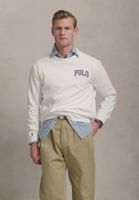 Balta džemperis su "Polo Ralph Lauren" tekstu, uždėtas ant mėlynai ir baltai dryžuoto marškinėlio, derinamas su chaki spalvos kelnėmis ir laikrodžiu.