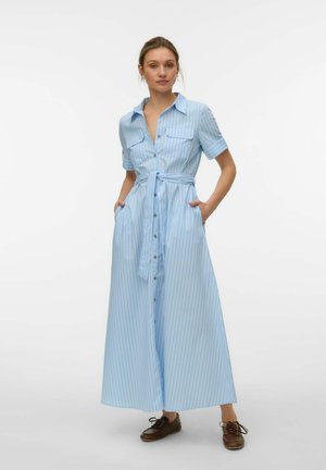 Kvinde iført en lyseblå, kortærmet, knaplukket maxikjole med tynde lodrette striber og taljebånd, kombineret med brune snøresko.
