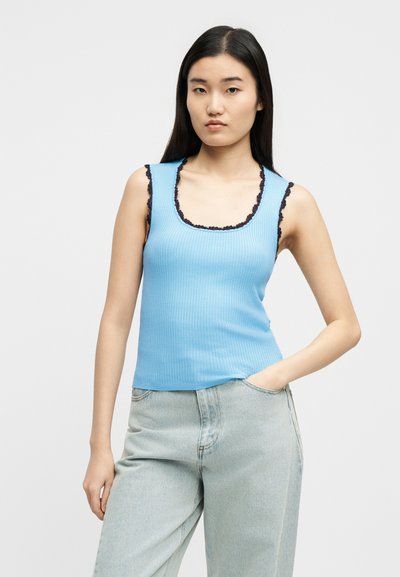 Νεαρή γυναίκα με μακριά σκουρόχρωμα μαλλιά φοράει γαλάζιο ριπ αμάνικο τοπ με μαύρη δαντέλα στο τελείωμα και τζιν ανοιχτού χρώματος, στέκεται με το ένα χέρι στην τσέπη.