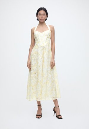 Kobieta w żółtej sukience midi na szyję z ażurowym wzorem i czarnych sandałach na wysokim obcasie, stojąca na jednolitym tle.