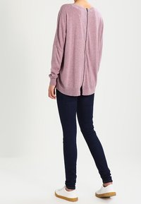 Jasnoróżowy sweter z długim rękawem, z zamkiem błyskawicznym z tyłu, o luźnym kroju i miękkiej teksturze, zestawiony z ciemnoniebieskimi skinny jeansami i białymi trampkami.