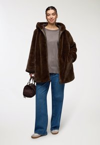 Cappotto con cappuccio in finta pelliccia marrone, top grigio, jeans a gamba larga blu, che tiene una piccola borsa marrone, dalla superficie liscia e vestibilità comoda.