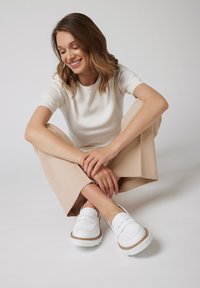 Białe mokasyny o teksturowanym wykończeniu, gumowej podeszwie i brązowych akcentach. Model ma na sobie kremową koszulkę i beżowe spodnie w szerokim kroju. Gładkie tło.