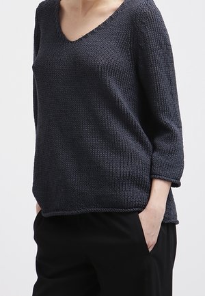 Kobieta ubrana w ciemnoszary dzianinowy sweter z dekoltem w serek i rękawami długości do łokcia, czarne spodnie, jedna ręka w kieszeni, na jednolitym tle.