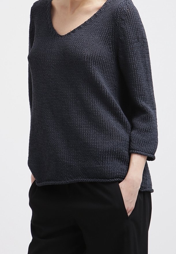 Femme portant un pull en tricot gris foncé à col en V avec des manches trois-quarts, un pantalon noir, une main dans la poche, sur un fond uni.