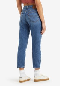 Personne portant un jean bleu taille haute à jambes droites, des chaussettes blanches et des baskets blanches, vue de dos avec un top court rose.