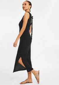Robe tricot noire avec une texture côtelée, présentant des fentes latérales, une silhouette ajustée et des liens en ruban au dos. Associée à des sandales plates.