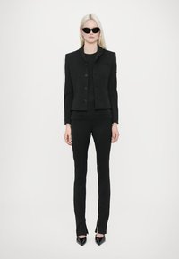 Black tailored blazer with buttons, paired with a fitted black top and trousers. Model wears black sunglasses and pointed-toe heels.