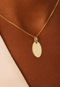 Gouden hangpendant aan een fijne ketting, met een ovale plaat met een gladde oppervlakte, hangend tegen een bruine huidtint achtergrond.