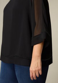 Czarna bluzka z przezroczystymi rękawami, ozdobiona falistym wzorem, o luźnym kroju. Dolny brzeg ma dekoracyjny wzór wycięć. Połączona z niebieskimi dżinsami.