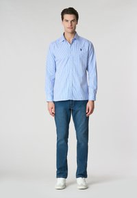 Camicia a quadri azzurra chiara con maniche lunghe, abbinata a jeans blu e sneakers bianche. La camicia presenta un piccolo logo sul petto.
