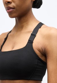 Κλειστό πλάνο γυναίκας που φοράει μαύρο αθλητικό σουτιέν με ρυθμιζόμενα τιράντες στους ώμους, σε λευκό φόντο.