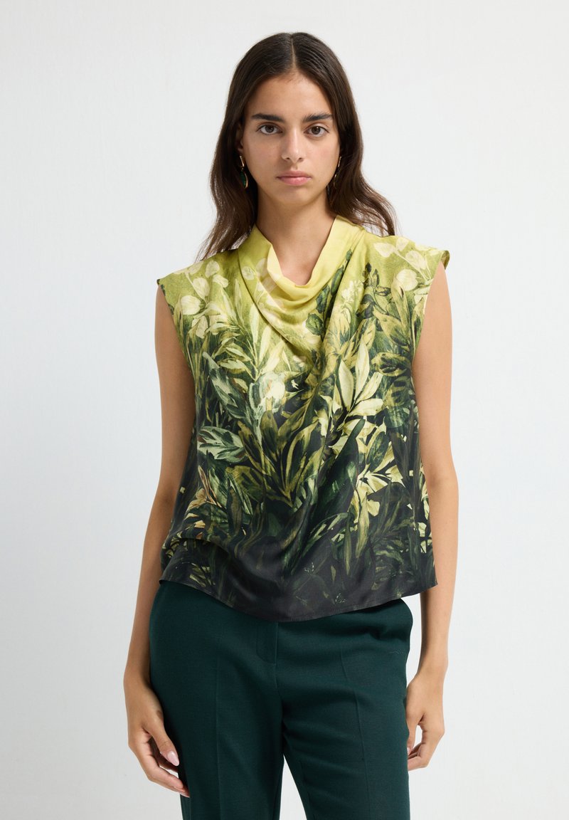 Vrouw in een mouwloos top met groene en gele bladerenprint en donkergroene broek, staande voor een effen witte achtergrond.