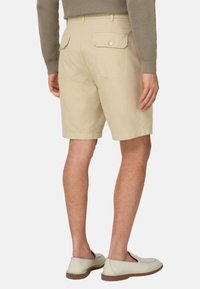 Homme portant un short beige à poches boutonnées au dos, un t-shirt à manches longues côtelé taupe, et des chaussures crème sans lacets, vu de dos.