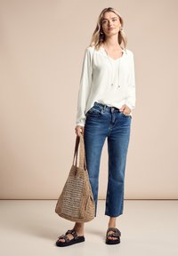 Witte blouse met lange mouwen en een verstelbare halslijn, blauwe recht gesneden jeans, geweven bruine schoudertas en zwarte sandalen met zilveren accenten.