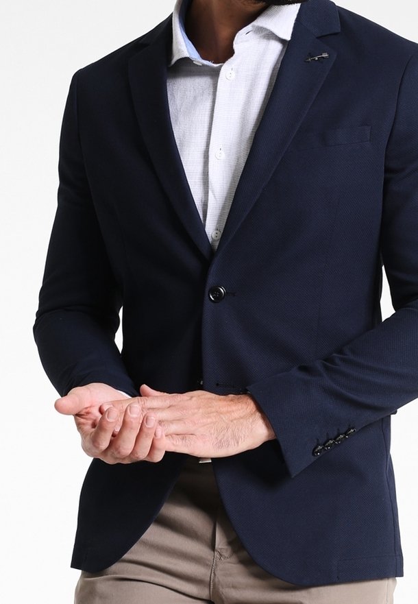 Marinblå blazer i texturerat tyg, med en enkel knappstängning och en subtil bröstfickdetalj, matchad med en ljus skjorta med krage under.