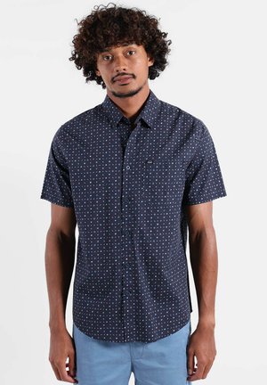 Homme aux cheveux bouclés portant une chemise à manches courtes bleu marine avec de petits motifs géométriques blancs et bleus, et un pantalon bleu clair.