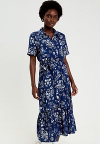 Mujer con vestido midi azul marino de manga corta con estampados blancos de flores y criaturas marinas, de pie con los brazos relajados frente a un fondo liso.
