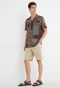 Camisa de manga corta con patrones geométricos multicolores en rojo, verde y negro, combinada con shorts beige y sandalias marrones.