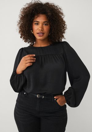 Woman with curly hair wearing a black long-sleeve blouse and black jeans, posing with one hand near her collar and the other in her pocket.