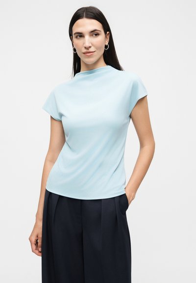 Γυναίκα με μακριά σκούρα μαλλιά που φορά ανοιχτό μπλε μπλούζα με κοντά μανίκια και μαύρο παντελόνι φαρδιάς γραμμής, στέκεται με το ένα χέρι στην τσέπη σε λευκό φόντο.