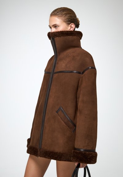 Naine kannab üle suuruse pruunist villast jope, millel on kõrge krae ja musta äärega, hoides musta käekotti valge tausta ees.