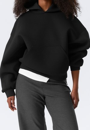 Persona con una sudadera negra de gran tamaño con bolsillo frontal, camisa blanca asomando y pantalones gris oscuro con fondo claro sencillo.