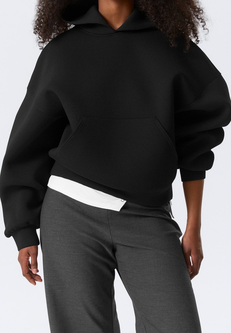 Personne portant un sweat à capuche noir oversize avec une poche devant, une chemise blanche qui dépasse, et un pantalon gris foncé sur un fond clair uni.