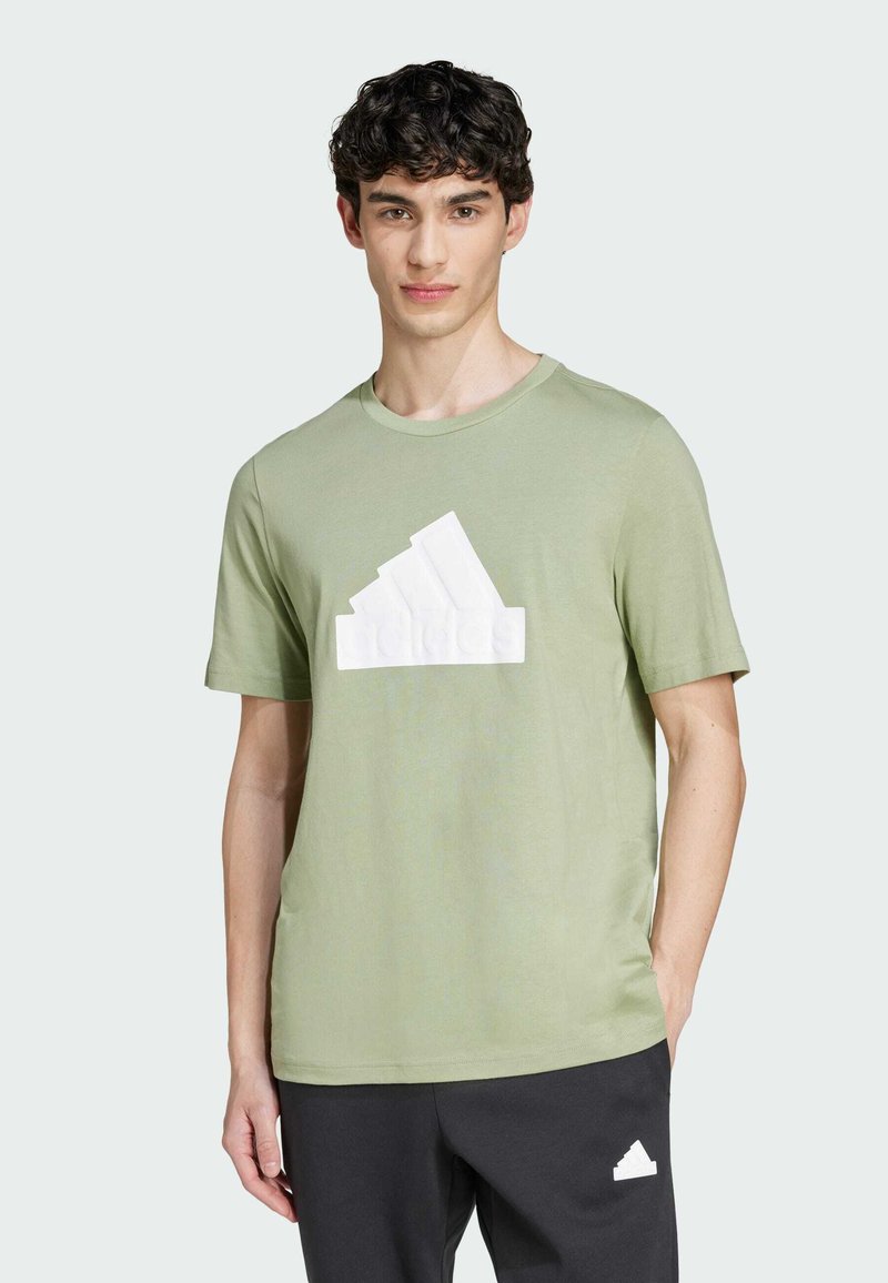 Camiseta de algodón verde claro con un diseño del logo de Adidas en relieve en blanco; mangas cortas y cuello redondo; combinada con pantalones gris oscuro.