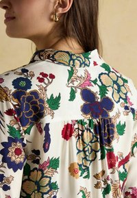 Floral-patterned blouse in cream with blue, green, pink, and burgundy flowers. Features a button-down collar and gathered back detailing.