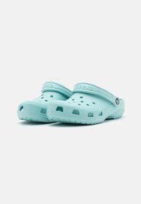 Clogs Crocs de cor azul claro, feitos de um material macio e durável, com orifícios de ventilação, uma sola grossa e a alça característica sobre o pé.