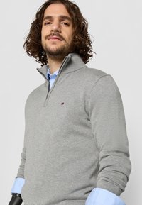 Pulover gri cu guler cu fermoar, completat cu o cămașă albastră deschis dedesubt. Prezintă un mic logo pe piept și manșete cu nervuri.