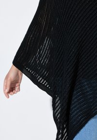 Svart stickad poncho med en öppen vävstruktur, utrustad med vida ärmar och en vågig kant, lagd över ljusblå denim.