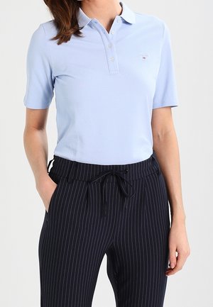 Mujer con una camiseta tipo polo de manga corta azul claro metida dentro de pantalones negros a rayas con cintura ajustable con cordón, con las manos en los bolsillos.