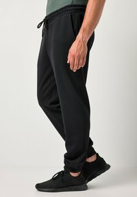 Mand iført sorte joggebukser med elastisk talje og sorte sneakers, stående mod en ensfarvet lys baggrund.