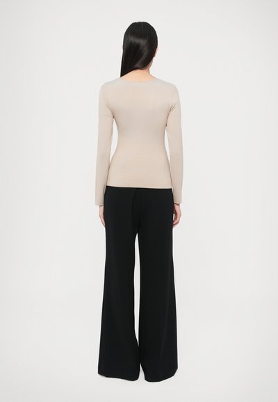 Nő háttal áll, bézs színű hosszú ujjú felsőt és fekete, bő szárú nadrágot visel, egyszínű fehér háttér előtt.