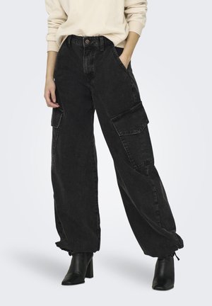 Pantaloni cargo neri in denim con vestibilità ampia, tasche laterali e caviglie elastiche. Indossati con stivali neri con tacco alto.