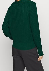 Mörkgrön stickad tröja med ett texturerat mönster, ribbade ärmslut och nederkant. Baksidan har en något loose fit och en rundad halsringning.