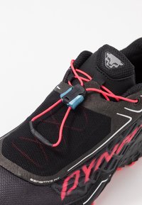 Černá atletická bota s texturovaným síťovinovým svrškem, červenými tkaničkami a modrým zapínacím mechanismem. Má vzorovanou podrážku a výrazné logo.