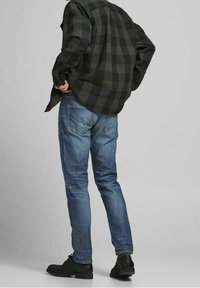 Camisa de franela de cuadros oscuros, jeans azules con bolsillos traseros y zapatos negros. Costuras visibles y un ajuste entallado en material de denim.